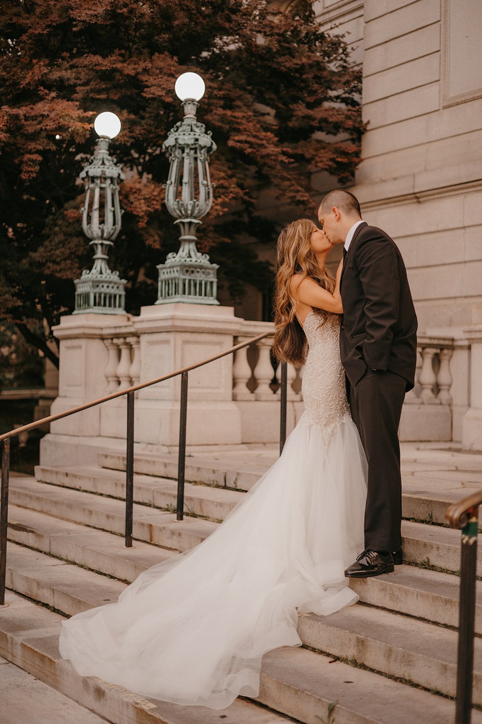 A bride in a mermaid style beaded wedding dress is kissing a groom in a black tuxedo on the stairs
