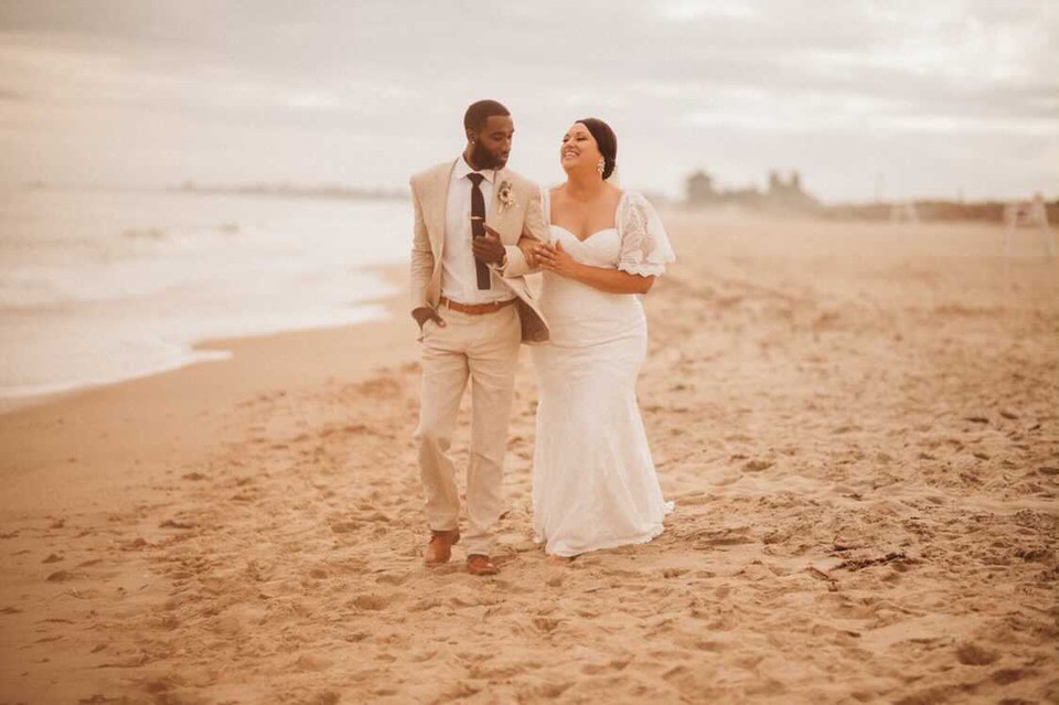 a bride and groom walking on the beach. The bride is wearing a wedding gown and the groom is wearing a suit
