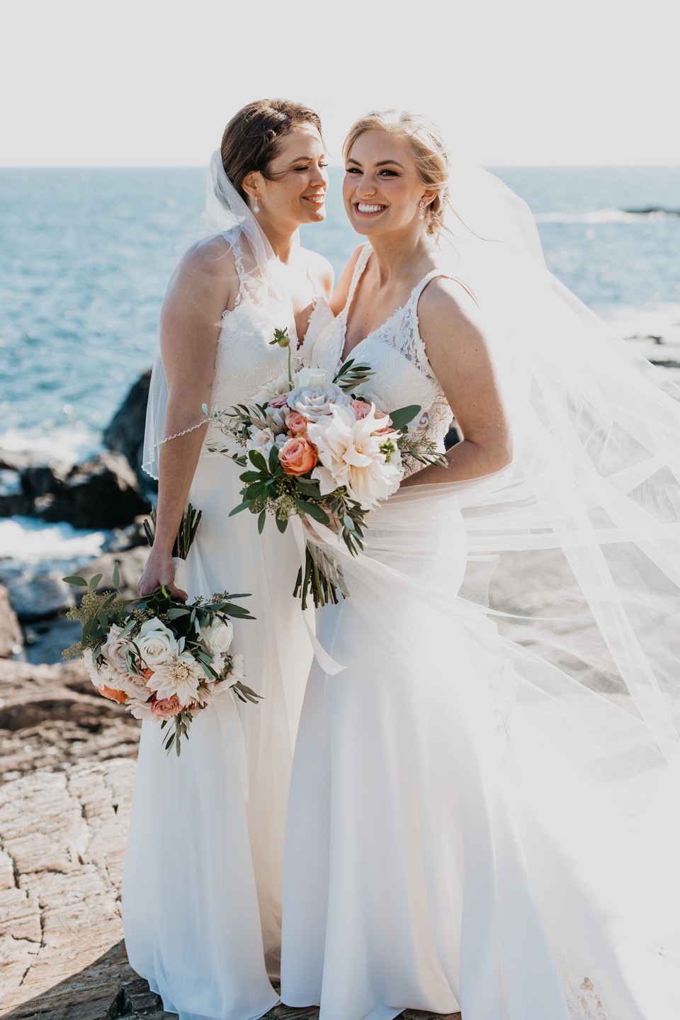 two brides wearing wedding dresses smiling on their wedding day by the ocean